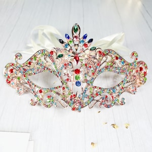 May include: A gold masquerade mask with a delicate, intricate design. The mask is adorned with colorful rhinestones and crystals, creating a dazzling and festive look.