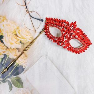 May include: A red jeweled masquerade mask with a gold stick handle. The mask is made of red rhinestones and has a delicate, intricate design.