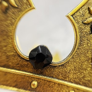 May include: Close-up of a gold-colored crown with a black gemstone. The crown has a detailed, textured design and a circular opening. The gemstone is faceted and sits prominently on the crown's surface.