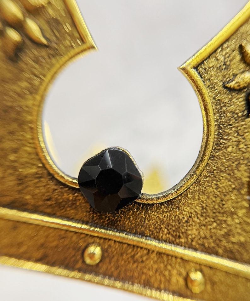 May include: Close-up of a gold-colored crown with a black gemstone. The crown has a detailed, textured design and a circular opening. The gemstone is faceted and sits prominently on the crown's surface.
