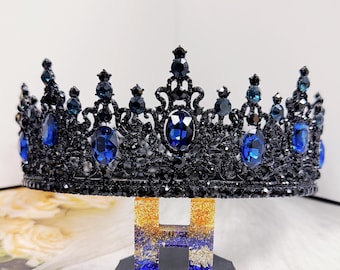 May include: A black crown with intricate filigree design, adorned with numerous blue and black gemstones. The crown is displayed on a decorative stand. The gemstones are oval and round, adding sparkle and elegance to the piece.