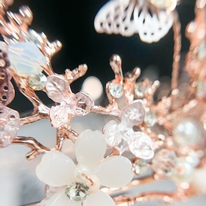 May include: Close-up of a rose gold floral crown with white flowers, clear crystals, and pearl accents. The crown features delicate branches and a butterfly design, perfect for weddings or special occasions.