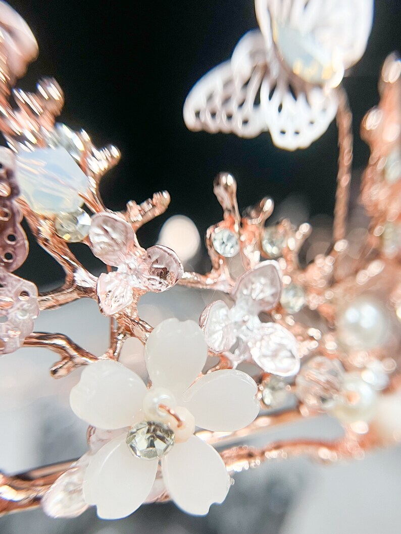 May include: Close-up of a rose gold floral crown with white flowers, clear crystals, and pearl accents. The crown features delicate branches and a butterfly design, perfect for weddings or special occasions.