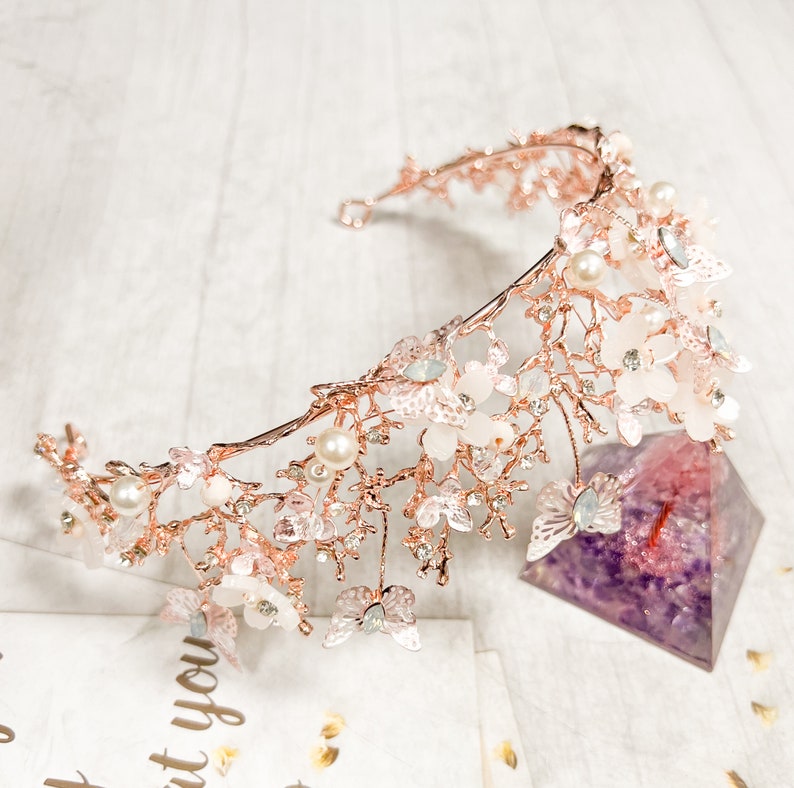 May include: A rose gold bridal crown with delicate floral and butterfly accents. The crown features faux pearls, clear crystals, and small white flowers. The design is intricate and elegant, suitable for weddings or special occasions.