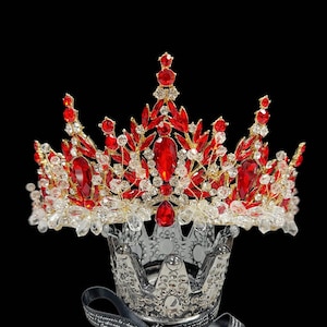 May include: A regal crown with a gold-tone frame, adorned with vibrant red and clear crystal-like gemstones. The crown is set against a black background, showcasing its intricate design and sparkling details. A smaller, clear crown is beneath it.