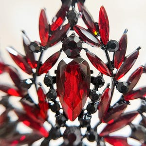 May include: Close-up of a decorative crown or tiara with a black metal frame. The crown is adorned with numerous red and black gemstones in various shapes, including teardrops and round accents. The central gem is a large, faceted red stone.