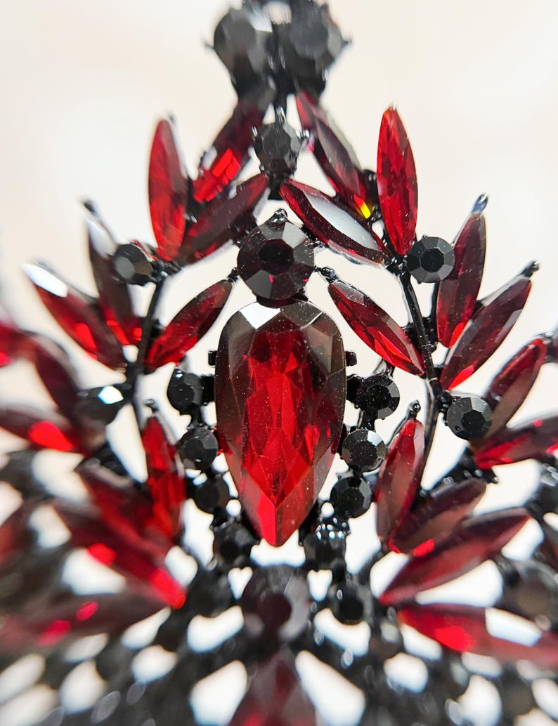 May include: Close-up of a decorative crown or tiara with a black metal frame. The crown is adorned with numerous red and black gemstones in various shapes, including teardrops and round accents. The central gem is a large, faceted red stone.