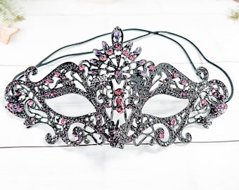 May include: A black masquerade mask with a delicate, intricate design. The mask is adorned with pink and purple rhinestones and has a black elastic band.