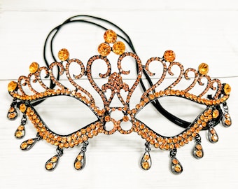 May include: A black masquerade mask with a crown design and orange rhinestones. The mask has a black elastic band and dangling rhinestones.