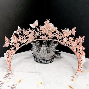 May include: A rose gold tiara with butterfly and floral accents. The crown features delicate butterflies and flowers, with small pearl-like embellishments. The tiara is set against a black background and a white surface.