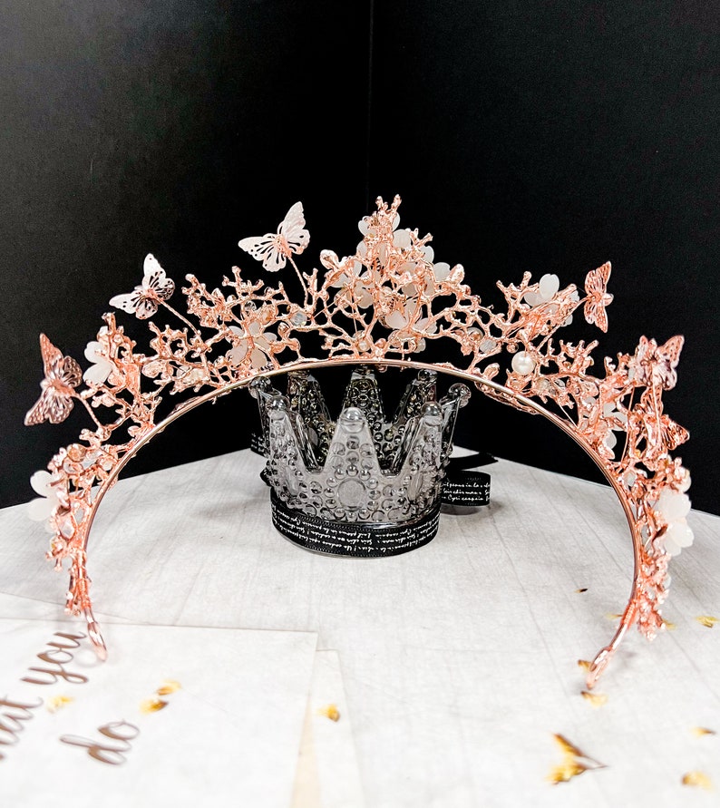 May include: A rose gold tiara with butterfly and floral accents. The crown features delicate butterflies and flowers, with small pearl-like embellishments. The tiara is set against a black background and a white surface.