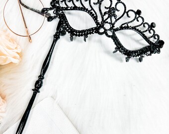 May include: A black masquerade mask with intricate detailing and rhinestones. The mask is attached to a long black stick.