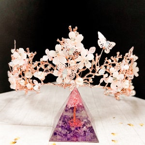 May include: A rose gold floral crown with white flowers, pearls, and butterfly accents. The crown sits atop a purple and pink pyramid-shaped object. The crown is designed for special occasions.