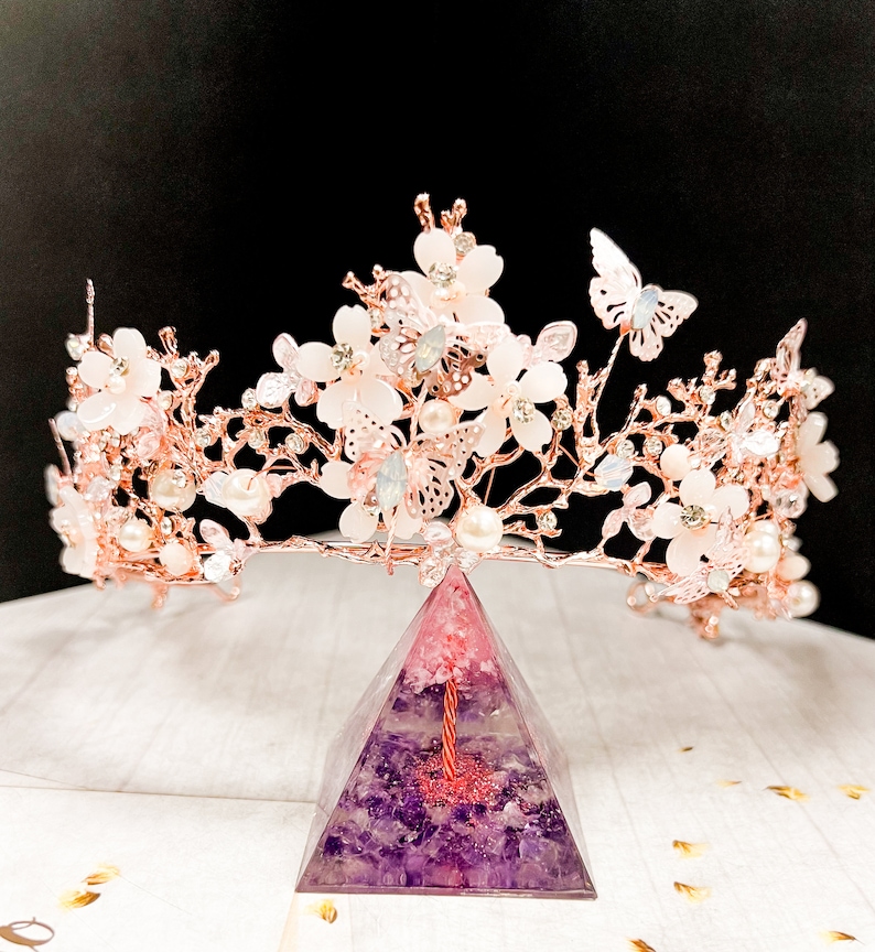 May include: A rose gold floral crown with white flowers, pearls, and butterfly accents. The crown sits atop a purple and pink pyramid-shaped object. The crown is designed for special occasions.