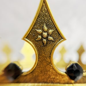 May include: Close-up of a gold-colored crown with a textured finish. The crown features a central, pointed element with a floral design and is adorned with black gemstones. The design suggests a regal or decorative theme.