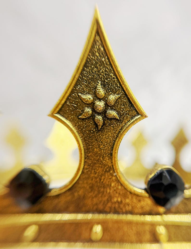 May include: Close-up of a gold-colored crown with a textured finish. The crown features a central, pointed element with a floral design and is adorned with black gemstones. The design suggests a regal or decorative theme.