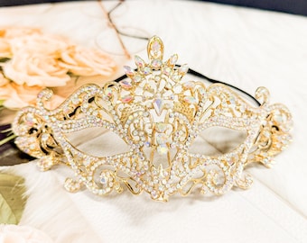 May include: A gold masquerade mask with a delicate, ornate design. The mask is covered in iridescent rhinestones and has a black elastic band.