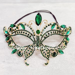 May include: A gold-toned masquerade mask adorned with emerald green rhinestones. The mask features a symmetrical design with a central jewel and a black elastic band for wear. The mask is designed for a formal event.