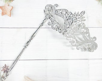 May include: A silver masquerade mask with an ornate, openwork design and a long handle. The mask is embellished with sparkling rhinestones and rests on a white wooden surface. The mask is a festive accessory for parties and events.