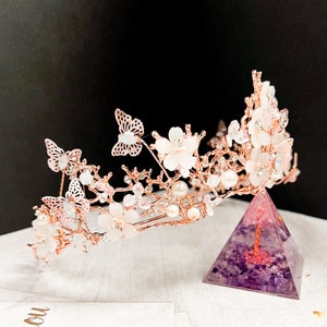 May include: A rose gold bridal crown featuring delicate butterflies, white flowers, and faux pearls. The crown is adorned with sparkling crystals and is designed for a wedding or special occasion. The crown is displayed on a purple pyramid.