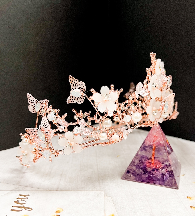 May include: A rose gold bridal crown featuring delicate butterflies, white flowers, and faux pearls. The crown is adorned with sparkling crystals and is designed for a wedding or special occasion. The crown is displayed on a purple pyramid.