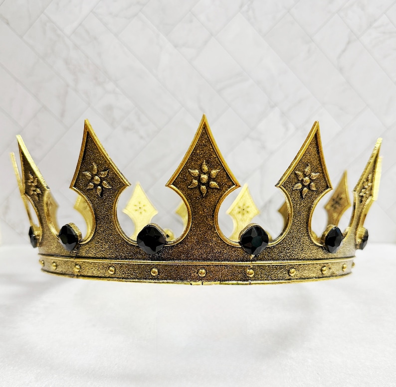 May include: A gold-colored crown with ornate details and black gemstones. The crown features pointed arches with floral designs and is likely for a costume or decorative purpose.