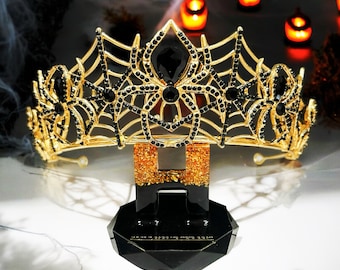 May include: A gold spiderweb crown with black rhinestones. The crown is sitting on a black and gold stand.