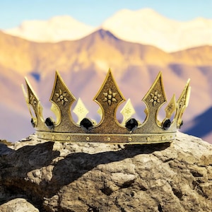 May include: A gold-colored crown with ornate details, including pointed arches and floral accents. The crown features black gemstones and sits atop a rock, with a mountain range in the background.
