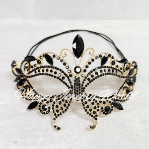 May include: A gold-toned masquerade mask with black gemstones. The mask features a butterfly-inspired design with a central teardrop-shaped gem and smaller gems outlining the eye and brow areas. A black elastic band is attached.