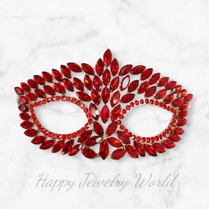 Puede incluir: Una máscara de carnaval roja con un diseño delicado hecho de pequeños diamantes de imitación brillantes. La máscara tiene la forma de una máscara veneciana tradicional con dos agujeros para los ojos y una parte superior puntiaguda.