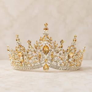 Könnte beinhalten: Eine metallische Krone mit einem Basisband aus klaren Strasssteinen und einem komplexen oberen Design aus bernsteinfarbenen tropfen- und marquiseförmigen Kristallen. Die Struktur umfasst mehrere vertikale Spitzen, die mit kleinen Steinen verziert sind.