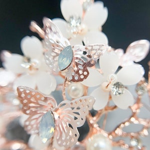 May include: A close-up of a bridal headpiece featuring white flower accents, rose gold branches, and delicate, openwork butterfly details. The headpiece also includes iridescent teardrop-shaped crystals and faux pearls.