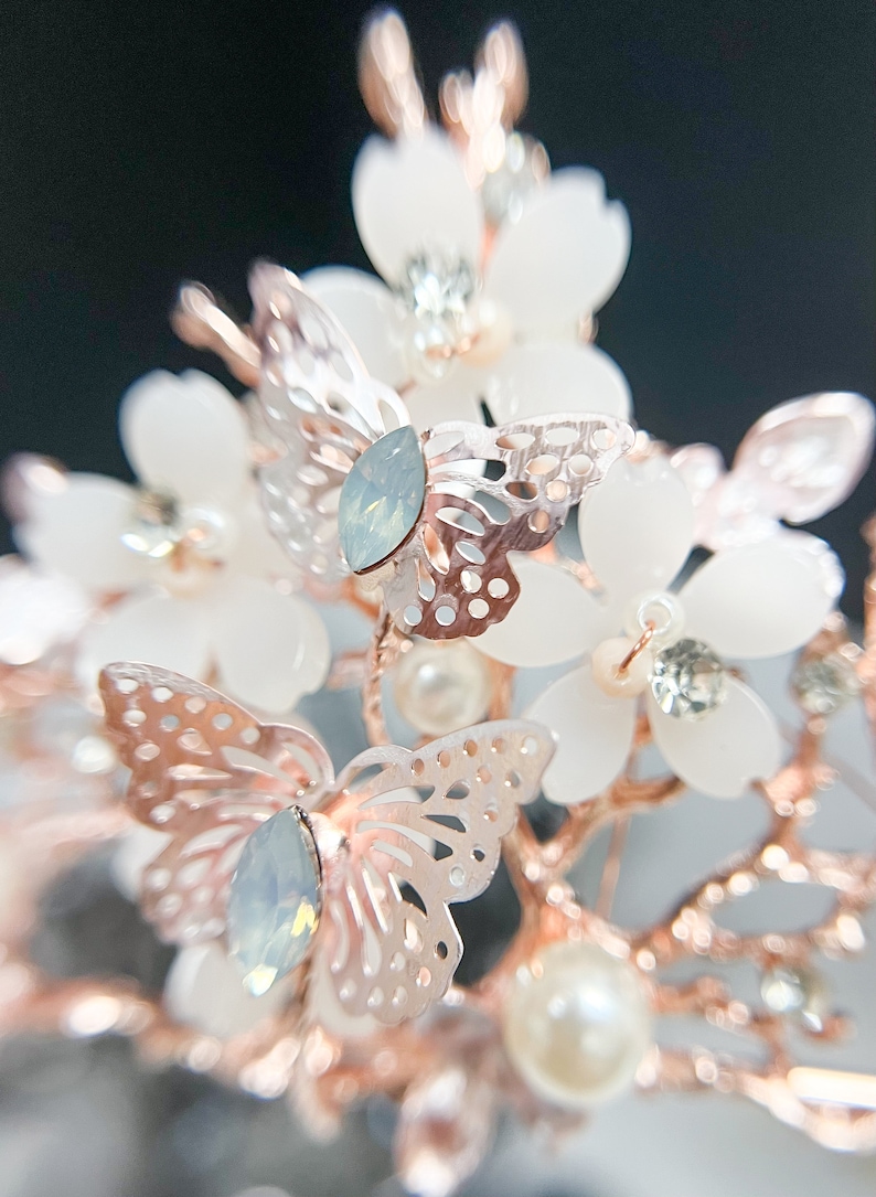 May include: A close-up of a bridal headpiece featuring white flower accents, rose gold branches, and delicate, openwork butterfly details. The headpiece also includes iridescent teardrop-shaped crystals and faux pearls.