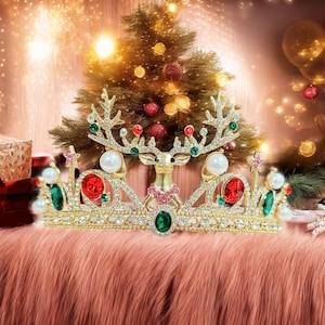 May include: A gold Christmas crown with a reindeer motif, embellished with red, green, and white gemstones. The crown includes pearl details and is set against a festive backdrop with a Christmas tree and twinkling lights.