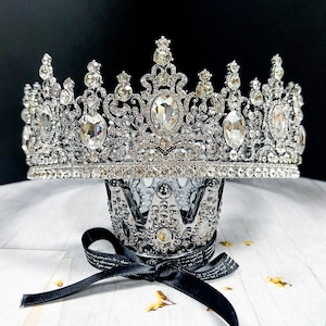 May include: An elaborate silver crown adorned with many clear gemstones. The crown features an intricate filigree pattern and sits on a smaller, matching crown, secured with a black ribbon. The background is a soft white surface against a dark backdrop.