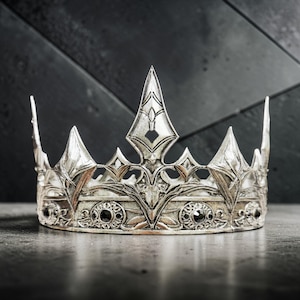 Könnte beinhalten: Eine silberfarbene Krone mit aufwendigen Details und mehreren Spitzen. Die Krone hat einen zentralen, hohen Dorn und ist mit schwarzen Edelsteinen verziert. Das Design deutet auf ein königliches oder Fantasy-Thema hin.