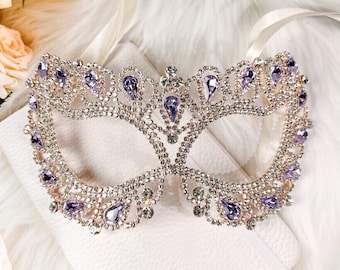 May include: A masquerade mask with a gold frame and purple rhinestones. The mask is decorated with a delicate pattern of small rhinestones.