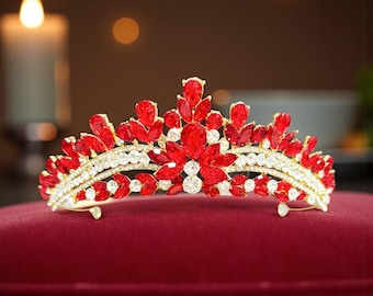 May include: A gold-toned tiara with red and clear gemstones. The crown features a floral design with large red stones and smaller clear crystals. The tiara is set on a red velvet surface, with a blurred background.