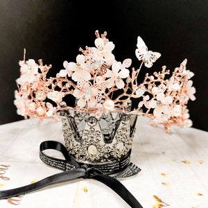 May include: A rose gold crown with delicate white flowers, pearls, and butterfly accents. The crown sits atop a silver decorative crown-shaped holder with a black ribbon tied around it. The crown is a beautiful accessory.