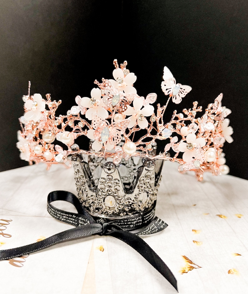 May include: A rose gold crown with delicate white flowers, pearls, and butterfly accents. The crown sits atop a silver decorative crown-shaped holder with a black ribbon tied around it. The crown is a beautiful accessory.