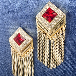 Peut inclure: Une paire de boucles d'oreilles dorées avec un strass rouge au centre de chaque boucle d'oreille. Les boucles d'oreilles sont de forme carrée avec une frange de chaînes dorées qui pendent du bas.