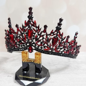 May include: A black and red jeweled crown with a teardrop-shaped pendant. The crown is adorned with red gemstones and black accents, displayed on a black stand with gold glitter accents. The base of the stand reads "YOU DESERVE THE REIGN."