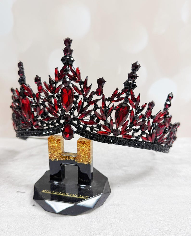 May include: A black and red jeweled crown with a teardrop-shaped pendant. The crown is adorned with red gemstones and black accents, displayed on a black stand with gold glitter accents. The base of the stand reads "YOU DESERVE THE REIGN."
