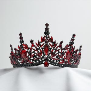 May include: A black and red jeweled crown with a symmetrical design. The crown features large, teardrop-shaped red gems and smaller red and black accents. The base is black, and the crown sits on a white surface.