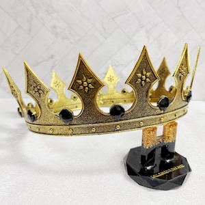 May include: A gold-colored crown with black accents and decorative floral patterns. The crown is displayed on a black stand with a geometric base. The crown features pointed arches and is adorned with black jewels.