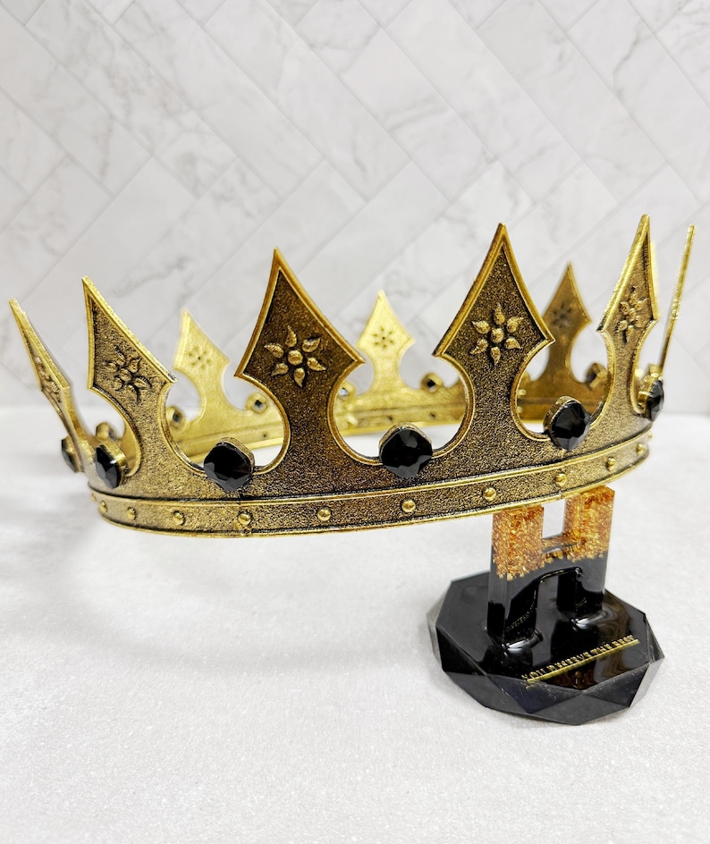 May include: A gold-colored crown with black accents and decorative floral patterns. The crown is displayed on a black stand with a geometric base. The crown features pointed arches and is adorned with black jewels.