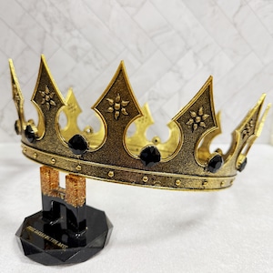 May include: A gold-colored crown with black gemstones, displayed on a black stand. The crown has ornate details and is likely a decorative item. The stand has text that reads "HOLD DAT... THE BEST".