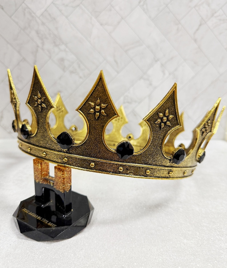 May include: A gold-colored crown with black gemstones, displayed on a black stand. The crown has ornate details and is likely a decorative item. The stand has text that reads "HOLD DAT... THE BEST".