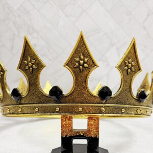 May include: A gold-colored crown with black accents. The crown features ornate detailing, including floral patterns and black gemstones. The crown is supported by a black and gold stand, perfect for a special occasion or costume.