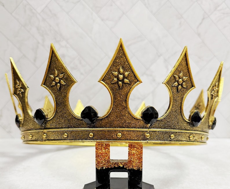 May include: A gold-colored crown with black accents. The crown features ornate detailing, including floral patterns and black gemstones. The crown is supported by a black and gold stand, perfect for a special occasion or costume.